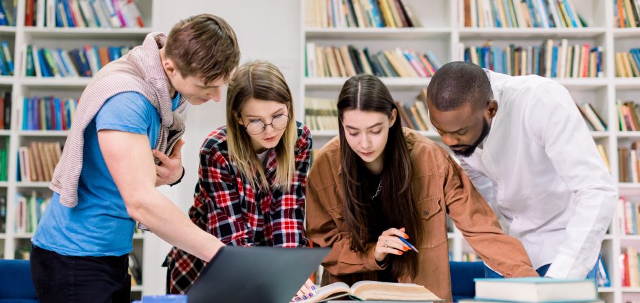 Front view of four concentrated multiethnic young people, college students preparing to their homework in library room, looking for needed information in book. Laptop and books on the table.