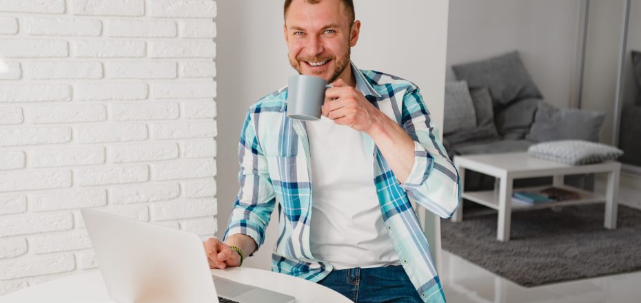 handsome smiling man in shirt sitting in kitchen at home at table working online on laptop from home freelancer, social distancing self isolation communication digital conference, remote worker