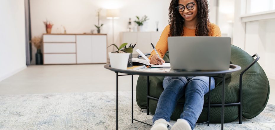 Check List. Smiling young black female in eyeglasses sitting on pouf chair at tea coffee table working on pc and writing letter in paper notebook, taking notes watching weninar, free copy space