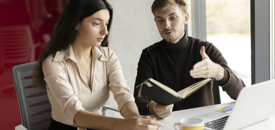 Workplace in a modern office. Attractive woman and man sitting at desk, guy explains to manager, holding notebook