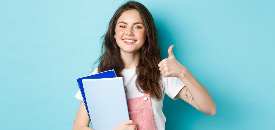 Young woman attend courses, girl student studying, holding notebooks and showing thumb up in approval, recommending company, standing over blue background.